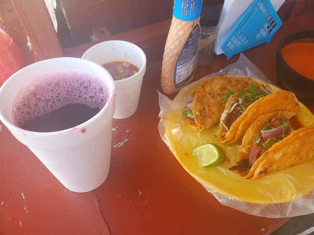 Archival photograph: birria tacos on a paper-lined plate with a cup of consomé and an agua fresca.
